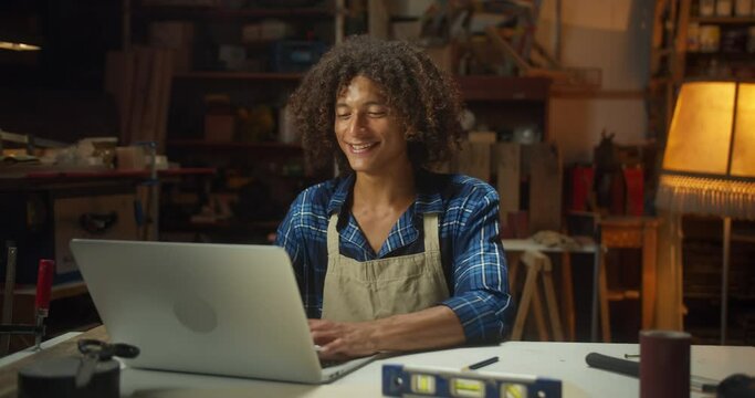 Happy curly Latin young man master in apron using laptop typing online in cozy workshop, handsome cheerful male carpenter smiling use computer on workbench staying up to date connected to internet