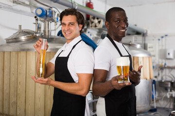 Young men brewers showing glass and flask of beer in brew-house