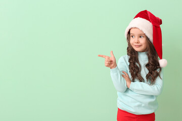 Fototapeta na wymiar Cute little girl in Santa hat pointing at something on color background