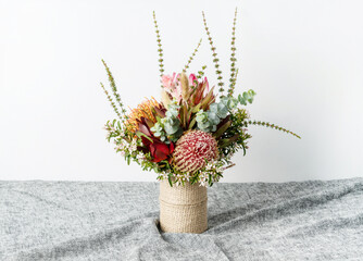 Beautiful Australian native flower arrangement of a red protea flower, orange and red banksias, red leucadendrons, white Eriostemons and eucalyptus leaves, in a rustic vase on a table.