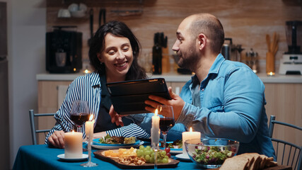 Fototapeta na wymiar Young caucasian husband using tablet during festive meal showing to his wife holiday destinations. Adults sitting at the table, browsing online, surfing, using smartphones, enjoying the meal