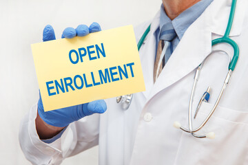 Close-up of a male doctor in gloves holding a sign with the text OPEN ENROLLMENT.