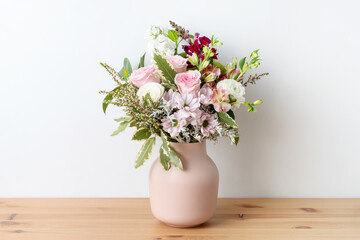 Beautiful cottage style flower arrangement, in a pink vase, on a wooden table. Flower bunch includes Roses, Snapdragons, Ranunculus, Daisy's, Chrysco, Lily of the Incas, Thryp and lush green foliage. 