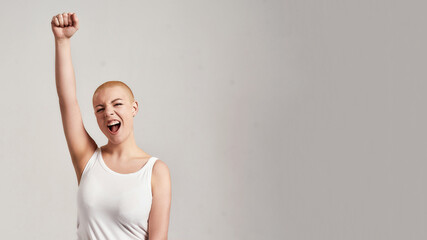 Portrait of a beautiful young caucasian woman with shaved head wearing white shirt, looking emotional, raising clenched fist, standing isolated over grey background