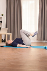 Elderly age woman doing abs physical training in living room on the yoga mat. Old person pensioner online internet exercise training at home sport activity with dumbbell, resistance band, swiss ball