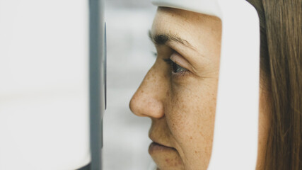 Close-up portrait of woman during test of refractometer machine, side view. Eye exam for young woman at optical store.