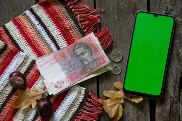phone with green screen and Ukrainian hryvnia and coins on a wooden table with oak leaf and chestnuts, autumn theme