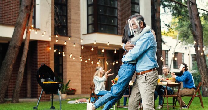 Happy Caucasian old grandfather in medical face mask hugging small granddaughter at court. Rear of little girl running to grandpa and embracing. Family in shields at barbeque meal on background.