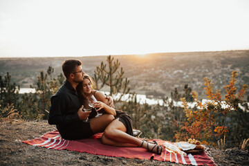 Naklejka na ściany i meble Young couple holding wine glasses while relaxing on picnic blanket on the hill at sunset. Happy couple toasting wine glasses. 