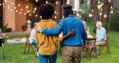 Caucasian man in face shield meeting young African American woman in yard and greeting with elbows. Middle-aged male inviting female for barbeque meal. Pandemic concept. Mixed-races people at picnic.