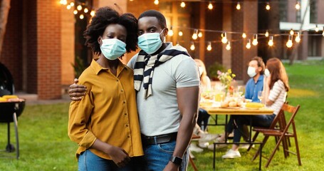 Portrait of African American young couple in medical mask standing in hugs in back yard. Caucasian people at table on background. Man and woman hugging at court in picnic or barbeque.