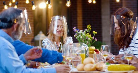 Happy mixed-races family in medical face shields sitting at table with meal outdoors at picnic and talking cheerfully. Joyful young and old people having dinner and speaking at barbrque. Coronavirus.