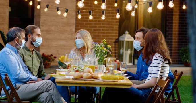 Happy Caucasian family in medical masks sitting at table with meal outdoor at picnic and talking. Joyful young and old people having dinner and having nice communication. Pandemic corona concept.