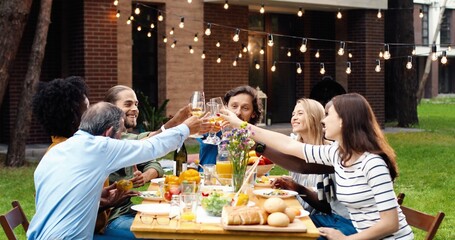 Happy multi ethnic family sitting at table with meal outdoor at picnic and toasting with juice and wine. Joyful mixed-races young and old people having dinner and toast at barbrque Weekend celebration