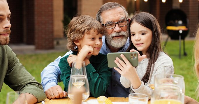 Happy multi ethnic family sitting at table with meal outdoor at picnic. Small kids sitting on grandfather knees and taking funny selfie photo on smartphone. Photos on mobile phone. Weekend Generations