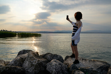 Woman's hands holding a smartphone and make photo during sunset on the lake. Travel photography taken a memory of the garda lake.