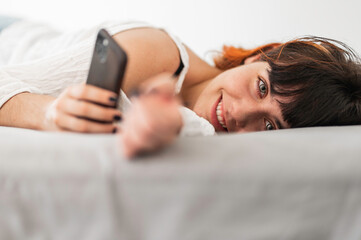 Obraz na płótnie Canvas Young woman with colored hair lying on bed and smiling while holding a smartphone in her hand