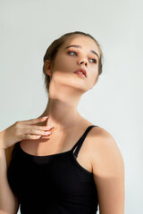 Fototapeta premium Aesthetic cosmetology. Beauty procedure. Young woman in black sleeveless top tenderly posing isolated on white in sun shine glow shadow. Skin care treatment. Female elegance