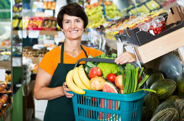 Adult female seller is demonstraiting basket with assortment goods in greengrocery