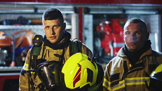 Portrait of two young firefighters on the rain in front of fire engine in full uniform. Fire dril. High quality 4k footage