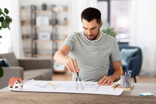 architecture, building, construction and real estate concept - male architect with compass measuring blueprint on table working at home office
