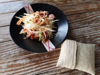 "Som Tum" green papaya salad in a plate eaten with sticky rice wrapped on a wooden table.