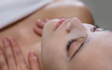 Young beautiful woman with white moisturizing fabric mask at her face in a beauty salon
