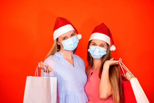 Female Friends Wearing Surgical Mask And Christmas Hat Holding Shopping Bags On A Vivid Red Orange Background