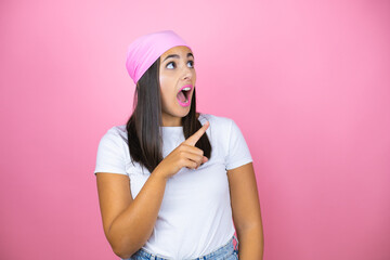 Young beautiful woman wearing pink headscarf over isolated pink background surprised and pointing with hand and finger to the side