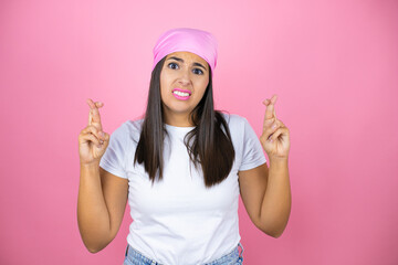 Young beautiful woman wearing pink headscarf over isolated pink background gesturing finger crossed smiling with hope and looking side