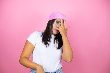 Young beautiful woman wearing pink headscarf over isolated pink background peeking in shock covering face and eyes with hand, looking through fingers with embarrassed expression