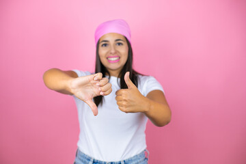 Young beautiful woman wearing pink headscarf over isolated pink background Doing thumbs up and down, disagreement and agreement expression. Crazy conflict