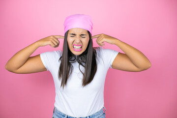 Young beautiful woman wearing pink headscarf over isolated pink background covering ears with fingers with annoyed expression for the noise of loud music. Deaf concept.
