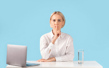 Attractive businesswoman making hush gesture at workplace near laptop computer, blue studio background