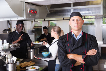 Experienced professional chef standing in kitchen of restaurant with working staff..