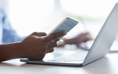 Unrecognizable black woman using smartphone and working on laptop in office, closeup