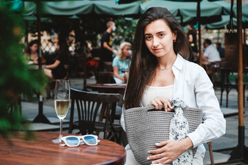 Beautiful young brunette woman wearing in sunglasses and white clothes, holding knitted bag sitting at the table of a street cafe. Copy, empty space for text