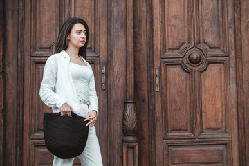 Beautiful young brunette woman wearing in white clothes, holding knitted bag on the background of old wooden doors. Copy, empty space for text