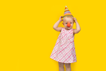 Little girl in party hat and red clown nose on yellow background. Childrens party. Copy space