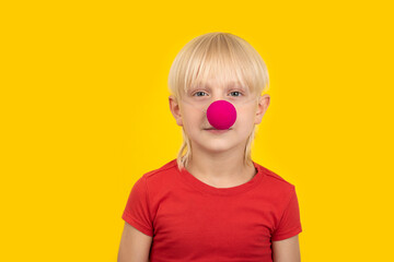Portrait of fair-haired boy with clown nose in red shirt. Yellow background.