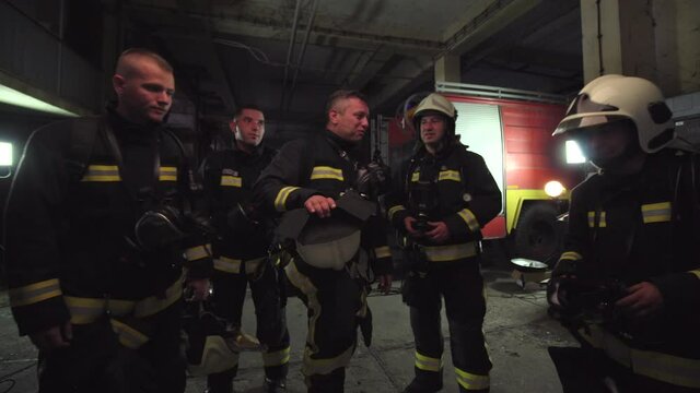 Group of five firefighters wearing full equipment in smokey area. Relaxed chatting after work well done. Fire trucks in the background. Name tag translation is fireman and model's last name.