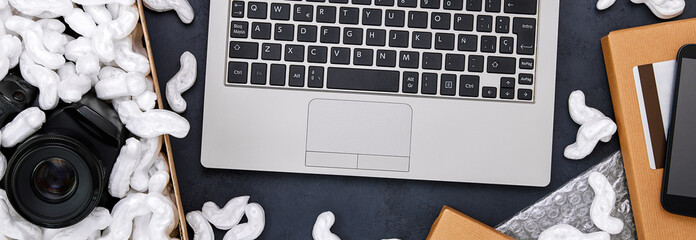 Online shopping concept on black web banner background. Laptop computer surrounded by smartphone, credit card, parcels and cardboard box with protective foam pads and electronic product inside.