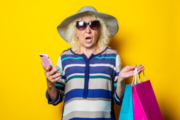 Old woman with surprised face in hat and glasses holds shopping bags with purchases and phone on yellow background