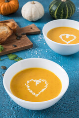 Two bowls of pumpkin soup with cream on blue background, decorated with little pumpkins, garlic and bread. Vertical stock photo.