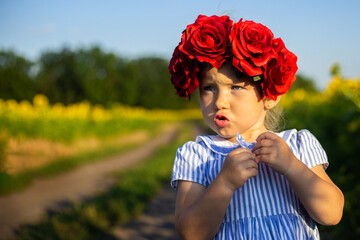Little girl in a dress and a wreath of large red flowers against the background of a sunflower field. Emotional face
