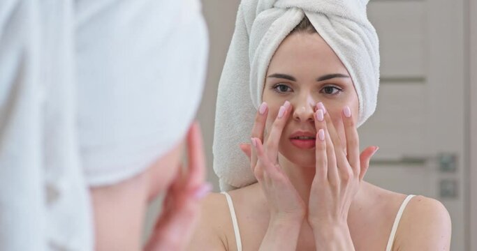 Young female stands in front of the mirror in her bathroom and applies cream on her face, skin care.