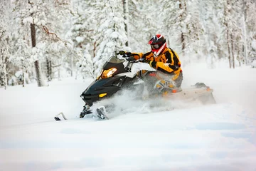 Fotobehang The racer in the outfit of a yellow-black overalls and a red-black helmet, driving a snowmobile at high speed riding through deep snow against the background of a snowy forest. © igor tsarev 