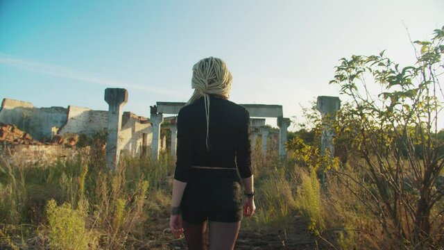 Rear view of slim attractive bloodthirsty female vampire walking through ruined building, turning back, showing her teeth and bloody face in rays of glowing sunset while looking for victim outdoors.