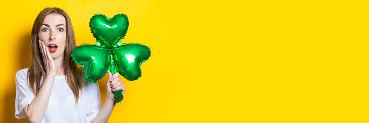 Young woman with a surprised face holds an air balloon in her hands a leaf of clover on a yellow background. Banner