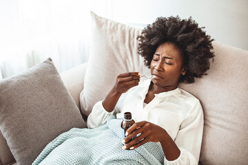 Woman taking cough syrup on sofa at home. Beautiful African young woman about to drink some cough...